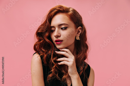 Curly lady with beautiful earrings poses in pink background. Red-haired wavy woman with beautiful makeup looking down on isolated backdrop..