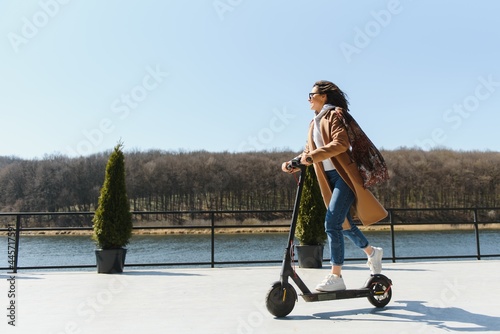 Young beautiful woman in a jacket smiles and rides an electric scooter to work along office buildings