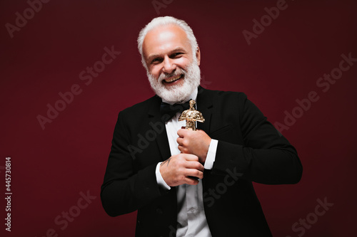 Cool man in black suit laughing and holding Oscar statuette. Smiling guy with grey beard and tattoos looking into camera..