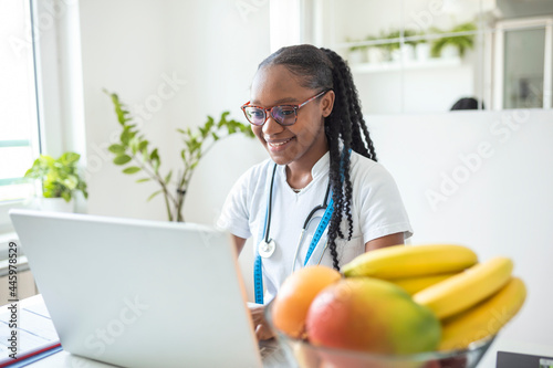 Portrait of young smiling female nutritionist in the consultation room. Nutritionist desk with healthy fruit, juice and measuring tape. Dietitian working on diet plan.