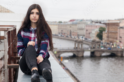 An Armenian girl with fluttering long black hair in a checkered shirt and jeans sits on the edge of a rooftop in the center of St. Petersburg on the Fontanka embankment