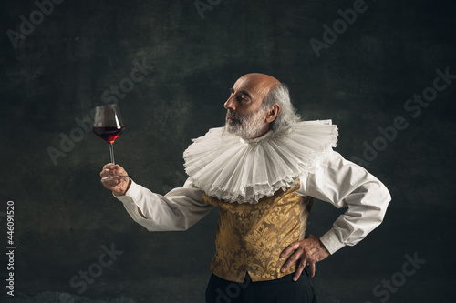 Elderly gray-haired man, medieval hystorical person, actor drinking wine isolated on dark vintage background. Retro style, comparison of eras concept.