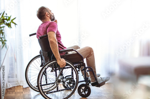 Handicapped person sitting in wheelchair and thinking about life, depression. Disabled mature man sitting on wheelchair and looking out the window in the living room at home