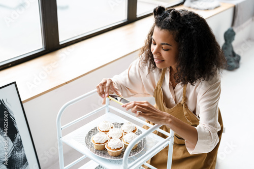 Black young woman in apron taking photo of her muffins at home