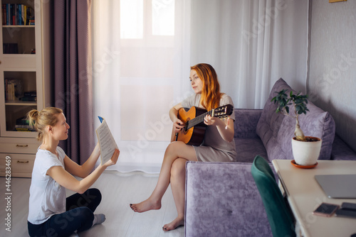 Young adult woman holds music notebook and looks at her friend playing the guitar. Two cute girls play music at home. Friendship, love, family leisure, hobbies.
