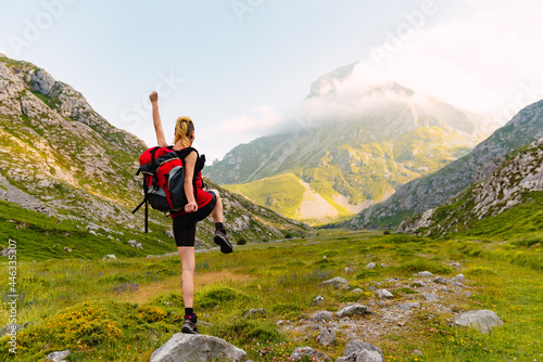 Caucasian and young person with his back turned, with a backpack and on top of a rock lifting one leg and one arm. Happy white woman hiking up the mountain