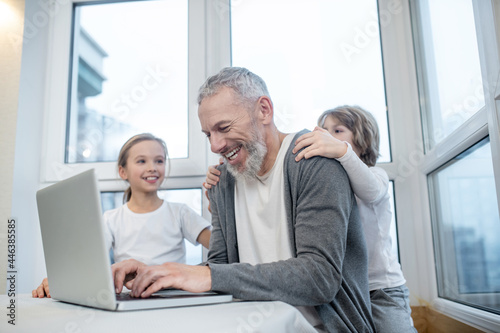 Gray-haired bearded man working on laptop while his kids interfering him