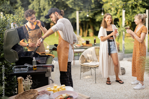 Happy young friends hanging out together, grilling food on a modern grill at beautiful backyard of a country house. People cooking food outdoors