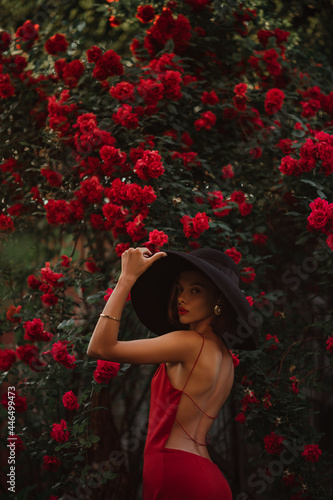 Elegant beautiful woman wearing luxury wide brim black hat, red dress with naked back, posing near blooming roses