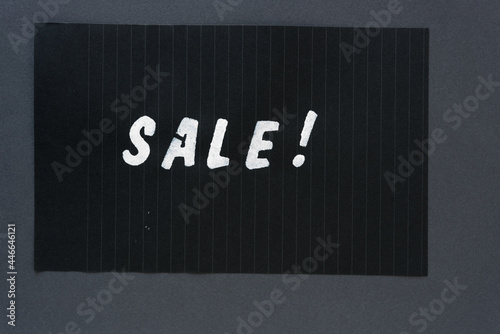 the word "sale!" in alpha/numeric plastic stencil letter type - hand painted in white acrylic paint - on black pad paper with faint lines - on dark gray paper