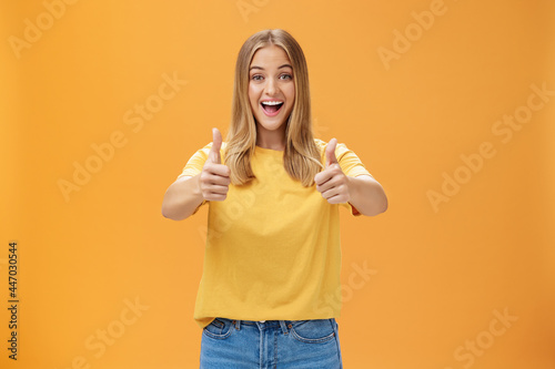 Woman supports with raised thumbs up and amused cheerful smile showing positive attitude expressing like on concept or idea giving approval posing happy and delighted against orange background