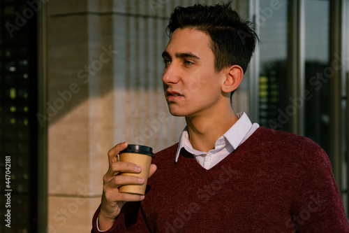 Handsome young man drinking coffee on his way to work. Businessman at lunchtime with a mug of tea. Walk along the autumn alley.