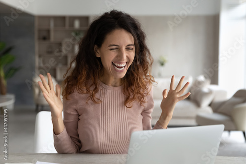 Overjoyed millennial Latino woman look at laptop screen feel euphoric read good news online. Smiling excited young Hispanic female triumph with promotion offer or discount sale deal on computer.