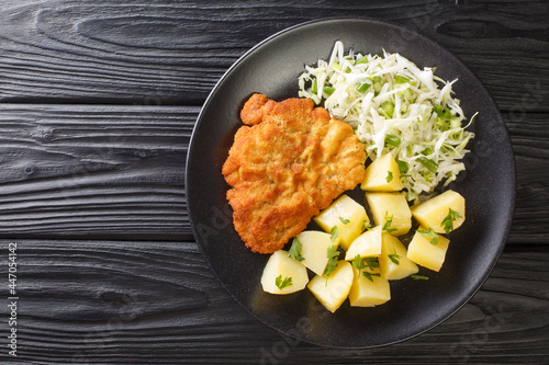 Kotlet Schabowy Polish Breaded Pork with boiled potatoes and cabbage salad close-up in a plate on a black wooden table. Horizontal top view from above