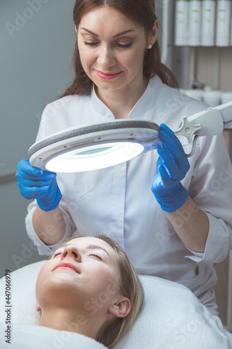 Beautician doctor is examining woman's face with special lamp