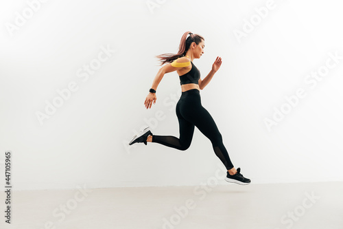 Runner in sport clothes sprinting near a white wall. Woman with kinesiology tape on shoulder jogging indoors.
