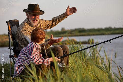 Grandfather and kid boy together fishing in the evening at sunset time in summer day on river in countryside, side view on caucasian multi-generation family, elderly man teach child boy to fish