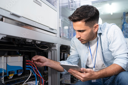 Young Caucasian male industrial engineer in blue shirt squatting touch and checking wire cables that is plugged on the back of the machine with the digital tablet in his hand in an industrial factory.