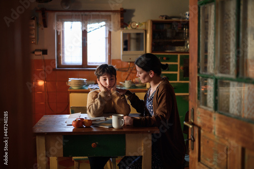 Poor mature mother and sad small daughter learning indoors at home, poverty concept.