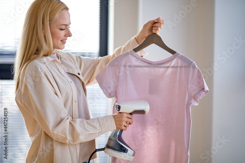 Woman using steaming iron to ironing casual t-shirt at home. Girl doing stream vapor iron for press clothes in hand. Blonde female doing household chores, housewife using smart device for ironing