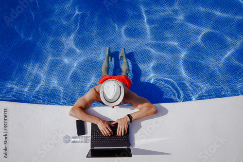 bird view of remote online working digital nomad man on workation with hat & laptop at a white table standing in a sunny turquoise water pool