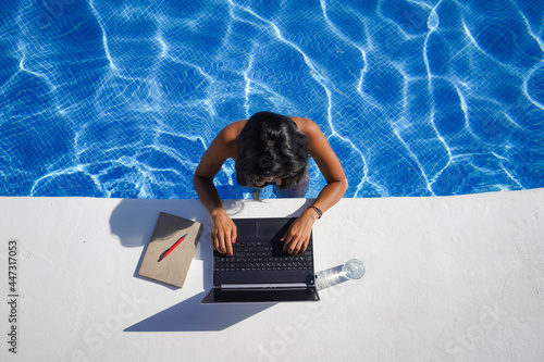 bird view of remote online working digital nomad woman in bikini with long black hair and laptop on a white table standing in a sunny blue water pool on Workation 