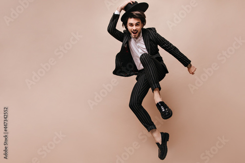 Attractive young brunette having fun and posing extraordinary against beige background. Man wearing white shirt, black striped suit, classic hat and elegant shoes