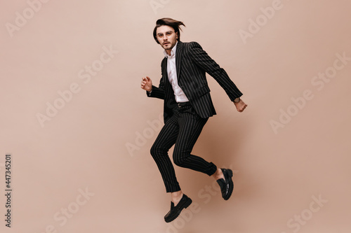 Full-length photo of jumping young man with brunette hair, glasses, white shirt and black suit. Model looking into camera and posing against beige background