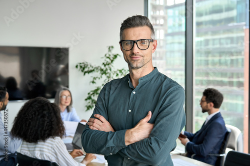 Smiling confident mature businessman leader looking at camera standing in office at team meeting. Male corporate leader ceo executive manager wearing glasses posing for business portrait arms folded.