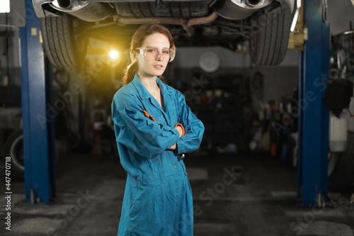 A young woman in work clothes, an apprentice stands next to the car and is proud and happy in the garage with protective goggles.
