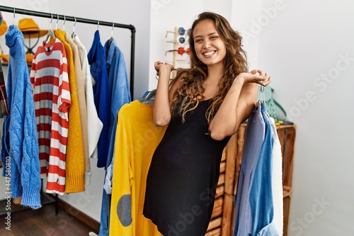 Young hispanic customer woman smiling happy holding hanger with clothes at clothing store.