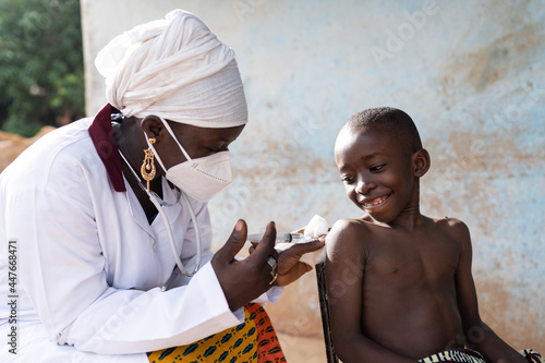 Black nurse with face mask injecting a dose of vaccine to a smiling brave schoolboy in an African hospital setting