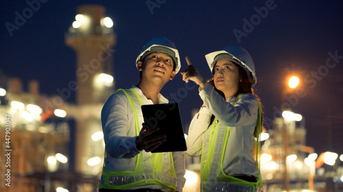 Two industrial engineer using digital tablet for work and discussing against the background of electrical power plant at night time.