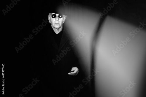 Spooky image of a man in black, wearing fedora hat, long coat and sunglasses in a dark place.