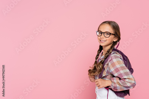 Preteen schoolgirl in eyeglasses and backpack smiling at camera isolated on pink