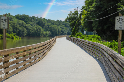 a long winding boardwalk along the river with a wooden rail along the sides with vast still river water, lush green trees with blue sky and a rainbow at Roswell Riverwalk Boardwalk in Roswell Georgia