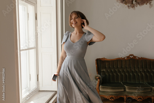 Pretty young lady posing at furnished studio. Cute short-haired brunette wearing maxi dress, smiling and moving with closed eyes near window against vintage sofa