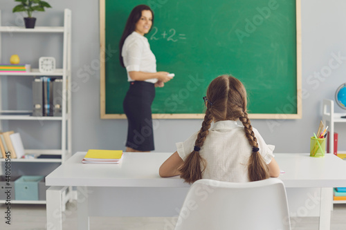 Pupil and teacher. Smart schoolgirl sitting at desk in modern classroom. Smiling tutor standing near blackboard waiting for her elementary student to do arithmetic sum. Primary education concept