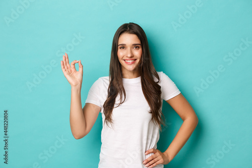 Image of confident smiling woman in white t-shirt, showing okay sign and looking pleased, agree or approve something good, praising excellent work, standing over blue background