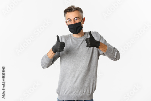Covid-19, pandemic and social distancing concept. Image of middle-aged man in black medical mask, gloves and glasses showing thumbs-up, protect himself from catching coronavirus