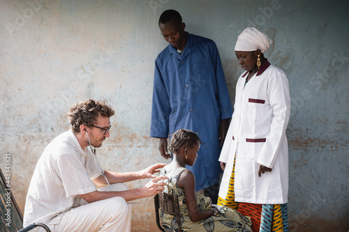 Two African physicians observing a young white doctor during auscultation of a small black girl's thorax during COVID screening
