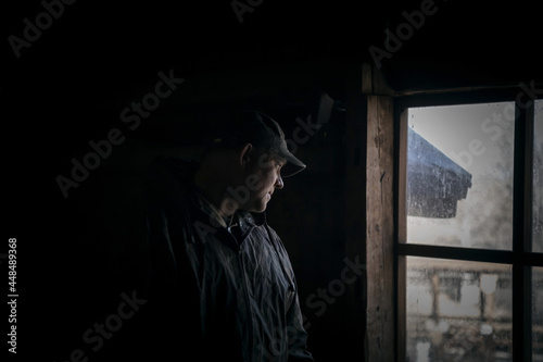 Silhouette of a suspicious strange man looking out the window in a scary dark attic of an old abandoned house in the darkness, thriller horror style
