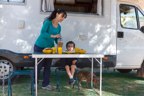 Mom serving food to her son in front of the motor-home, at the table in the shade of the tree.