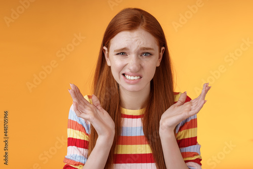 Awkward unhappy worried young redhead girl cringe feel sorry apologizing smirking smiling nervously frowning squinting spread hands sideways shrugging confused, standing orange background