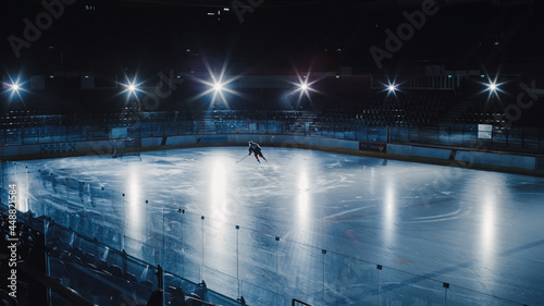 Ice Hockey Rink Arena: Professional Player Training Alone. Skates, Dribbles with Stick, Shooting, Hitting the Puck. Determined Athlete with Desire to Win, Be Champion. Cinematic Lights