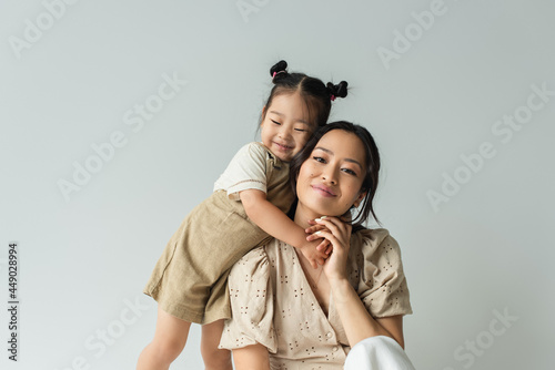 cheerful asian toddler daughter hugging happy mother isolated on gray