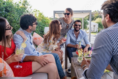 A group of laughing young people gathers at the aperitif party on the terrace. Millennials congregated in the garden joke together.