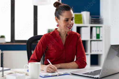 Smiling businesswoman reading data from laptop screen and taking notes on clipboard. Manager doing administrative wwork. Business leader doing financial expertise for client.