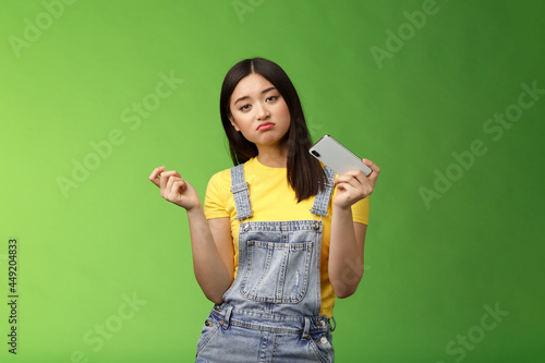 Bored indifferent cool urban asian girl waste time summer holiday alone, playing smartphone game, interrupt texting, browsing internet, gesturing reluctant, look sad, losing battle, green background