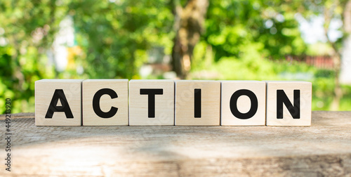 Six wooden blocks lie on a wooden table against the backdrop of a summer garden and create the word ACTION.